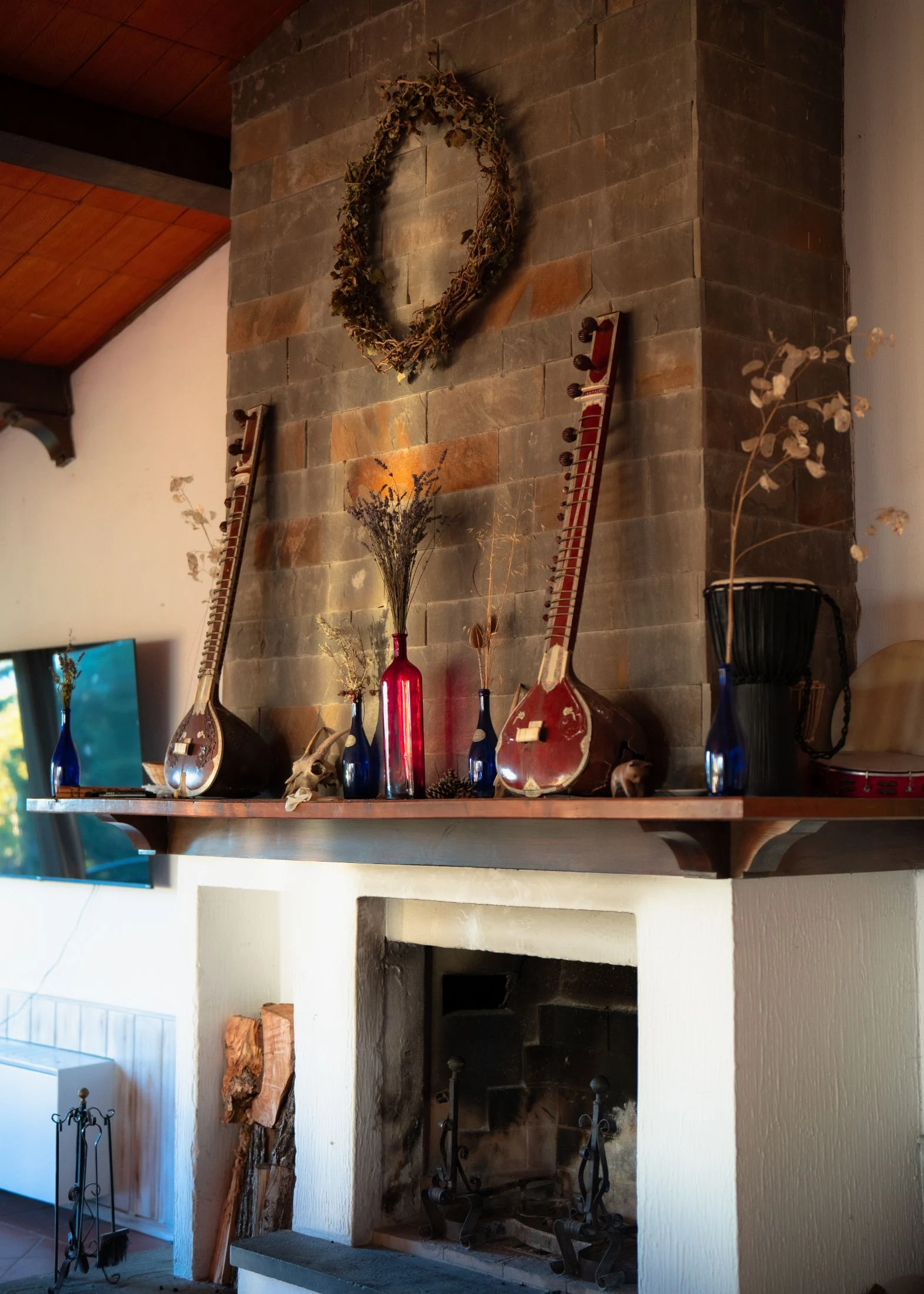 A rustic stone fireplace mantel at Maiolo displaying two sitar-like string instruments, a dried wreath, glass bottles with dried flowers, and a small djembe drum