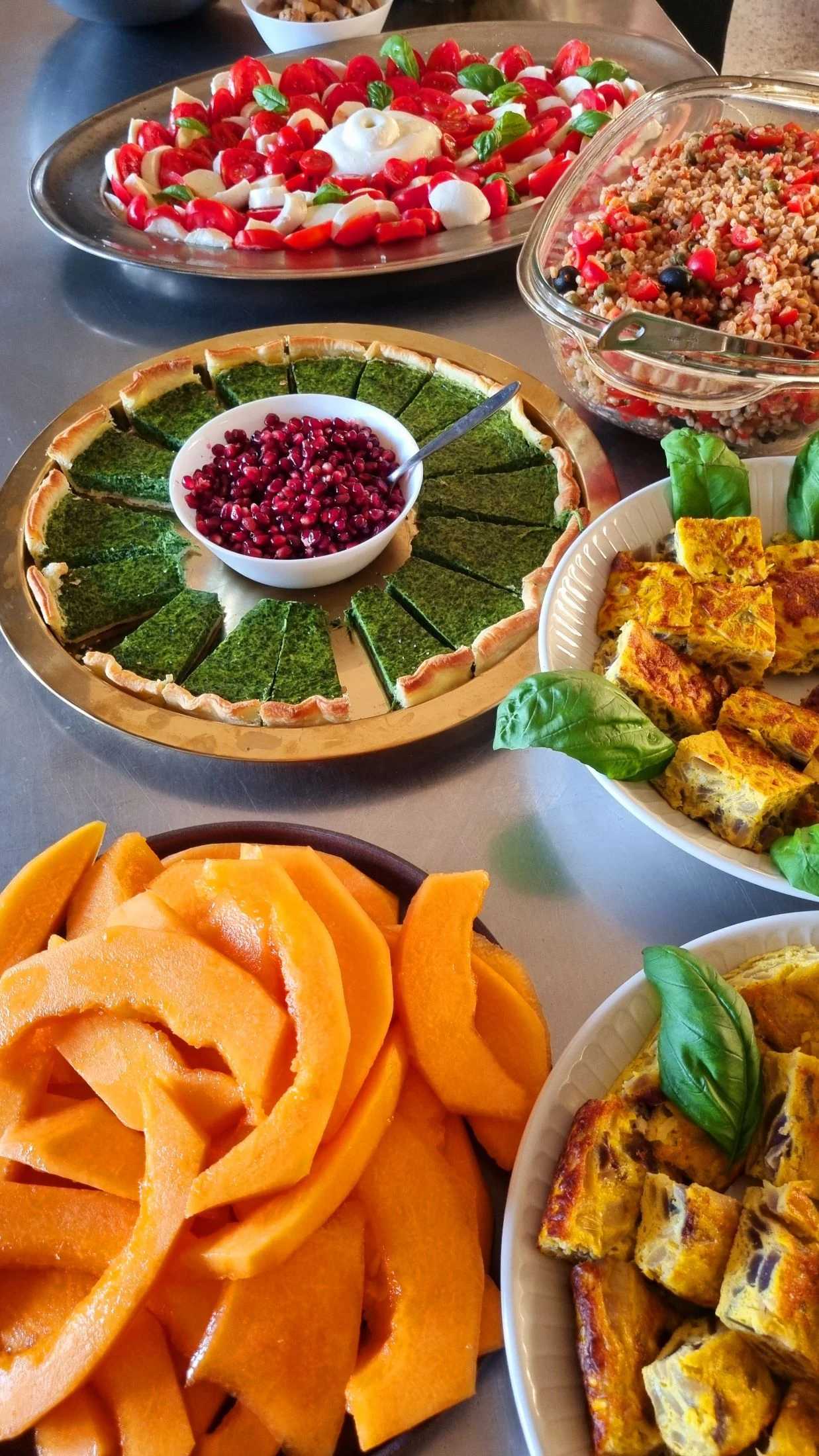 An overhead shot of a shared breakfast table with caprese salad, farro salad, a sliced spinach tart with pomegranate seeds, frittata squares, and cantaloupe