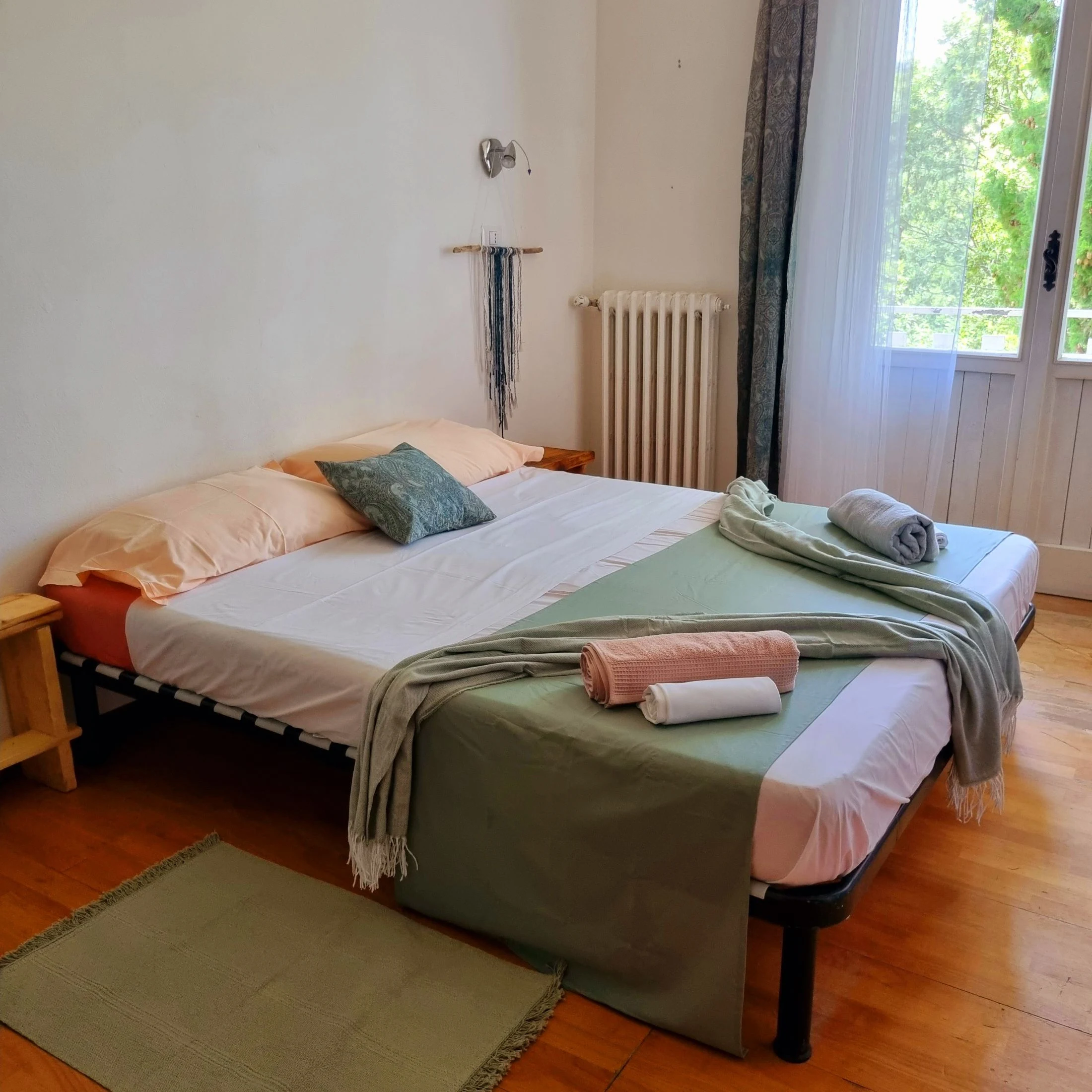 A made-up double bed with peach, white, and sage-green linens, folded towels, and a woven wall hanging above the headboard