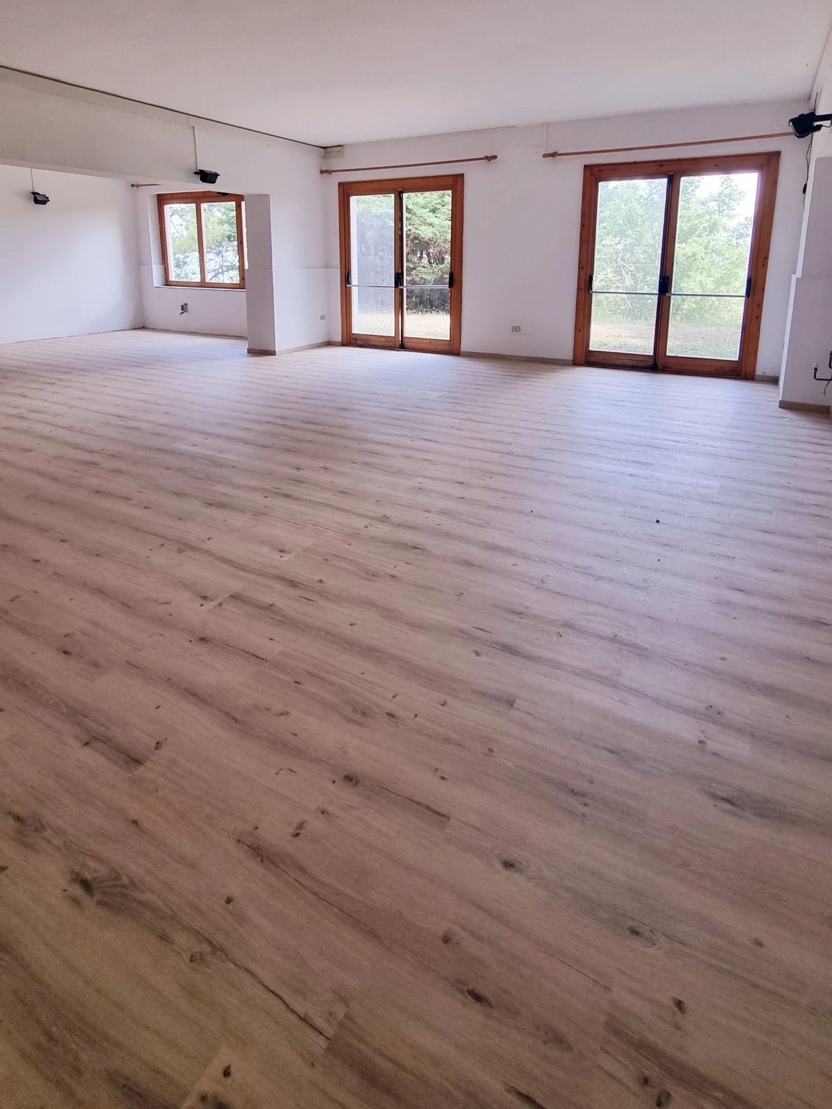 A spacious, empty room mid-renovation with fresh light wood-look flooring, white walls, and wood-framed glass doors opening onto greenery