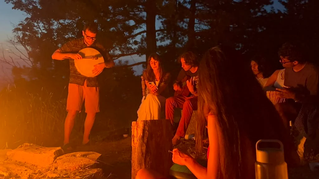 A group gathers on a firelit hillside at dusk, a man playing a banjo while others play guitar and hand drums nearby, warmly lit by the glow of a fire