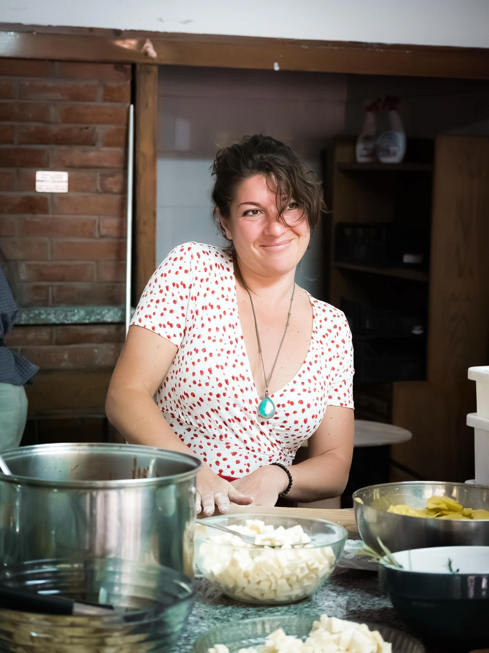 A woman smiles while standing at a kitchen counter behind bowls of diced ingredients, in a rustic brick-walled kitchen