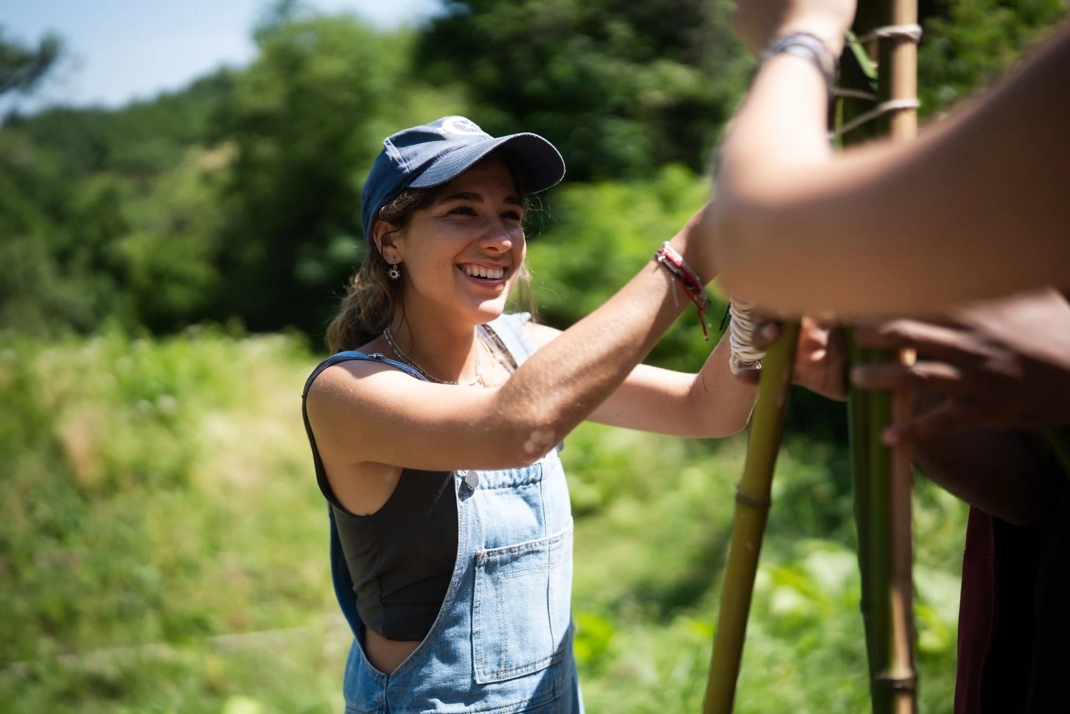 A woman in denim overalls helps another person lash bamboo poles together outdoors, green hills behind them, during a work block at Maiolo
