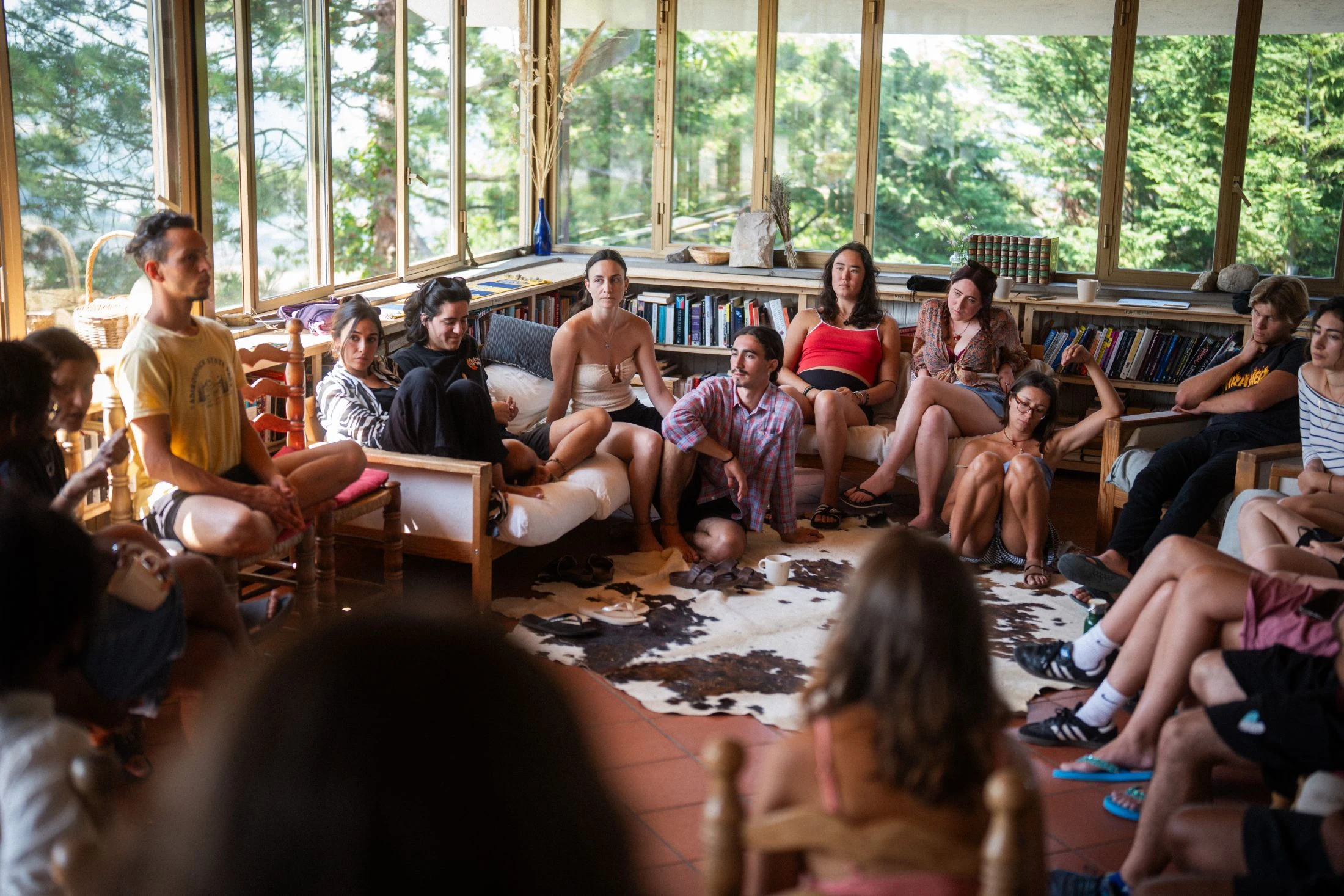 A large group of people sit in a circle on chairs, a sofa, and a rug in a bright window-lined sunroom at Maiolo, gathered in discussion