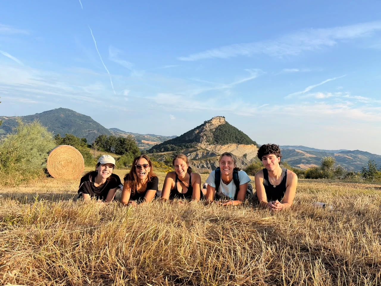 Five friends lie together in a golden harvested field beside a hay bale, laughing, with a distinctive rocky peak and layered hills behind them