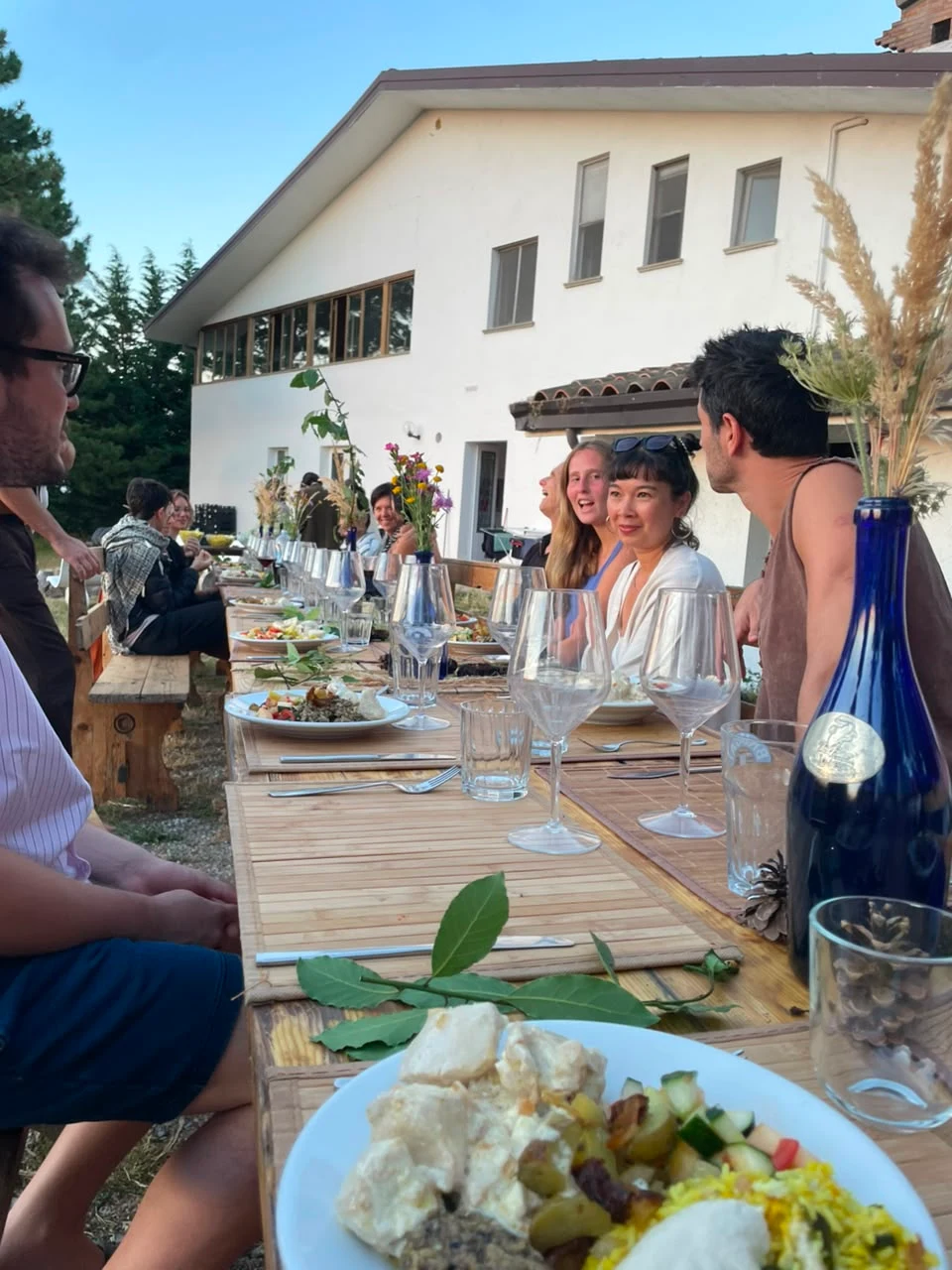 A long outdoor table set for a communal Shabbat dinner in front of the Maiolo farmhouse, guests laughing over wine glasses, plated food, and wildflowers at dusk