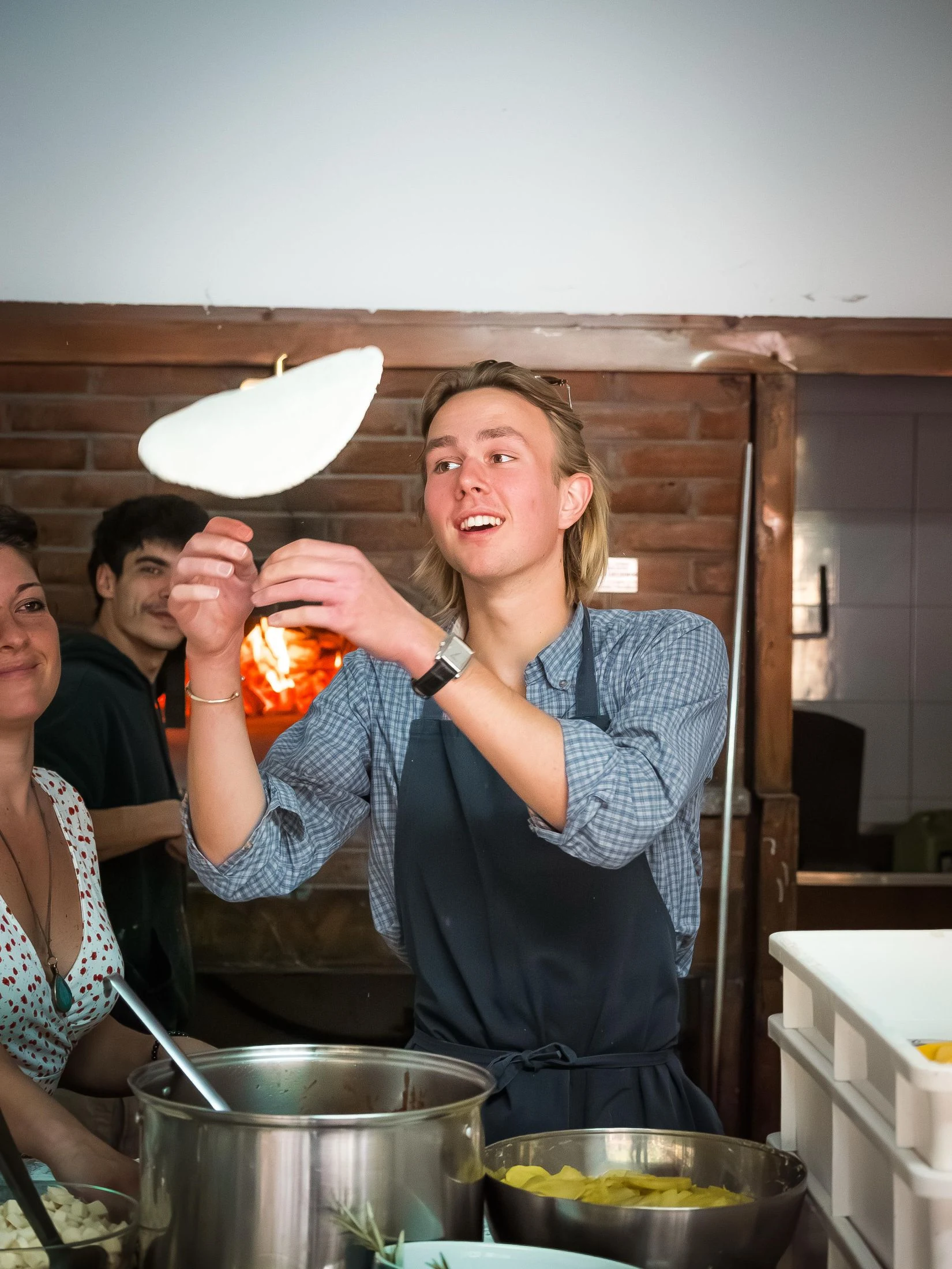 A young man in an apron tosses a flattened ball of pizza dough in the air in front of a glowing wood-fired brick oven