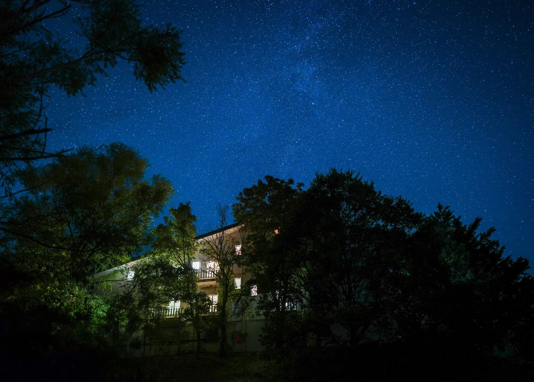 A night sky full of stars stretches above the large white farmhouse at Maiolo, its windows glowing warmly, silhouetted by dark treetops