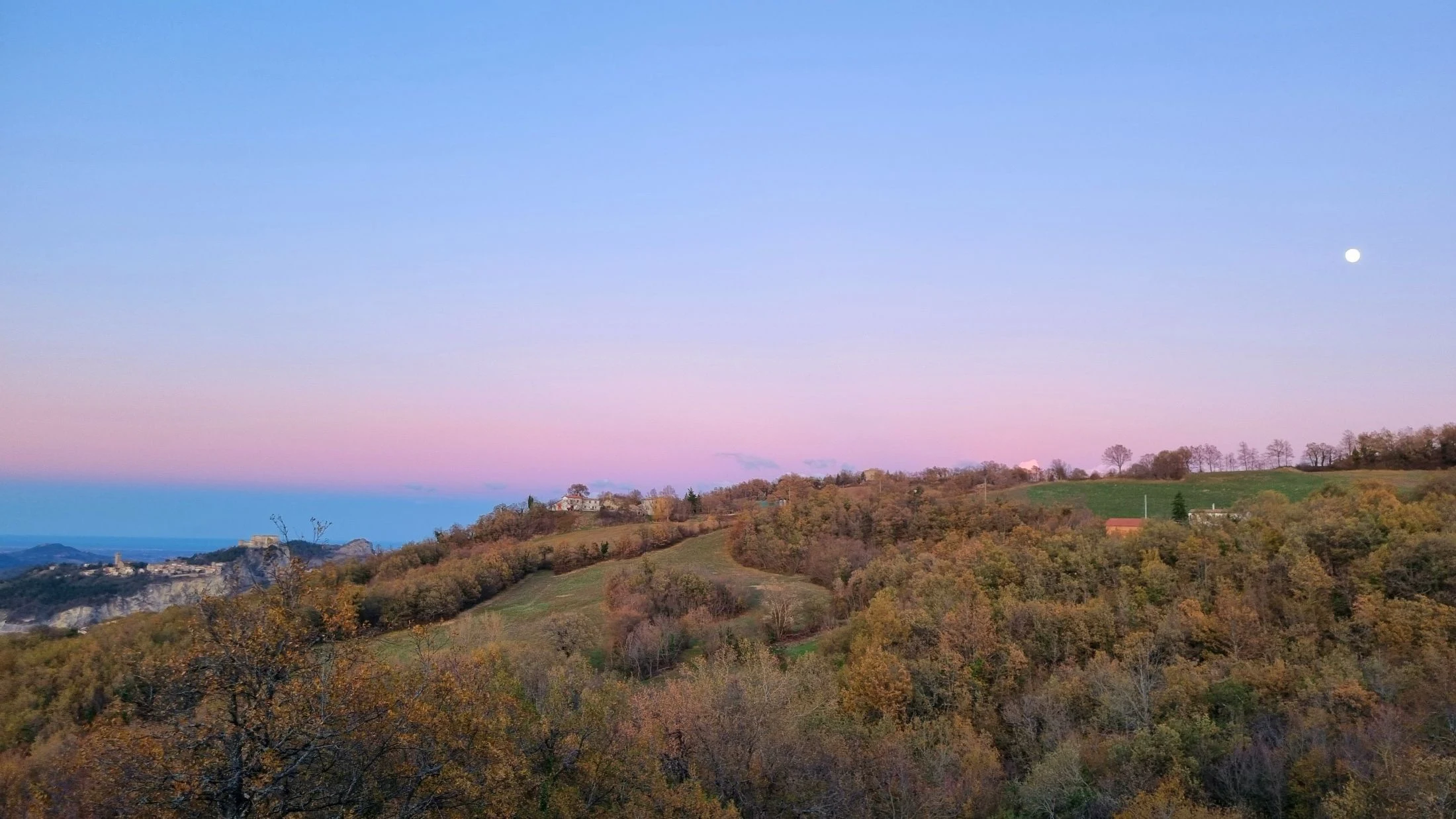 A wide view over rolling hills and a distant hilltop village at dusk, a moon visible in the pink-to-blue sky