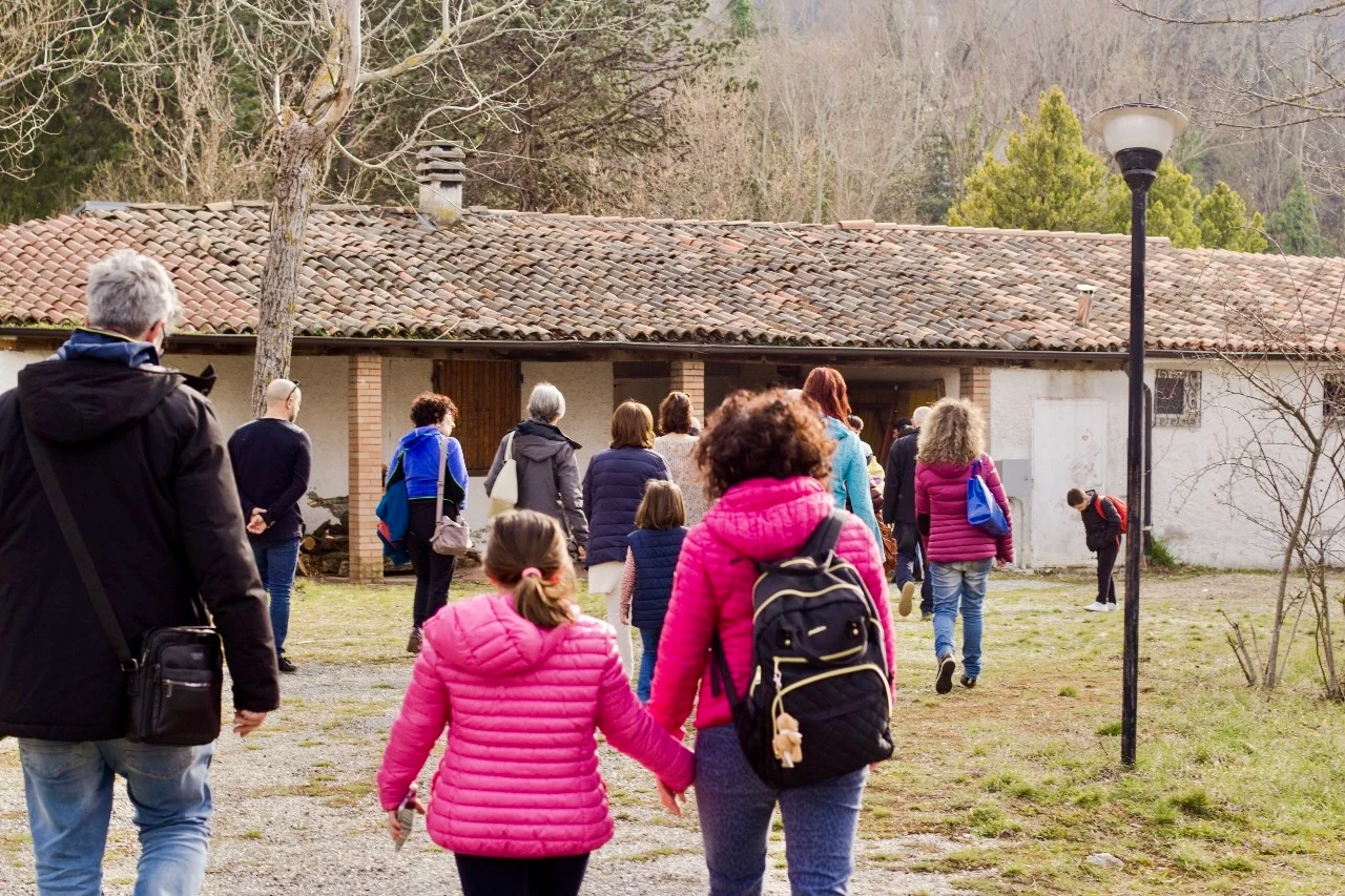 A group of people, including two children in matching jackets holding hands, walks across a grassy yard toward a long single-story farmhouse