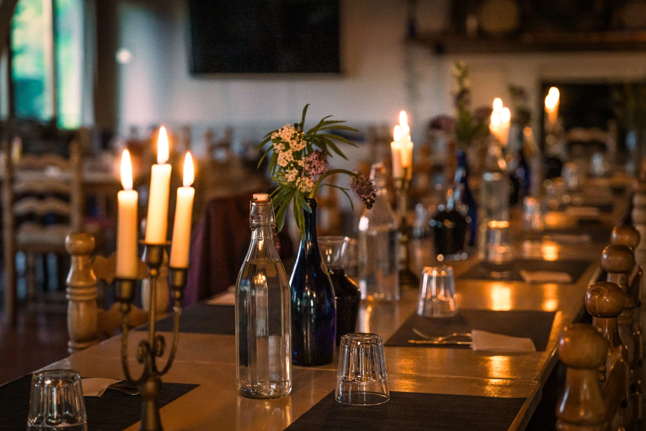 A long communal dining table set with brass candelabras, lit candles, glass water bottles, and small vases of wildflowers, stretching into a warmly lit dining room