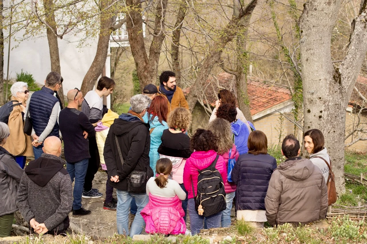 A mixed-age group stands and sits together outdoors near a stone building, listening to a man speaking to the group