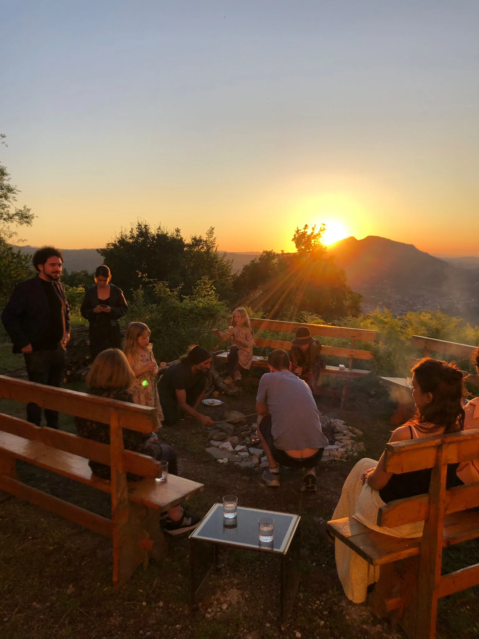A group of adults and children gathers on wooden benches around a small stone firepit at golden hour, a hazy sun setting over a hillside town