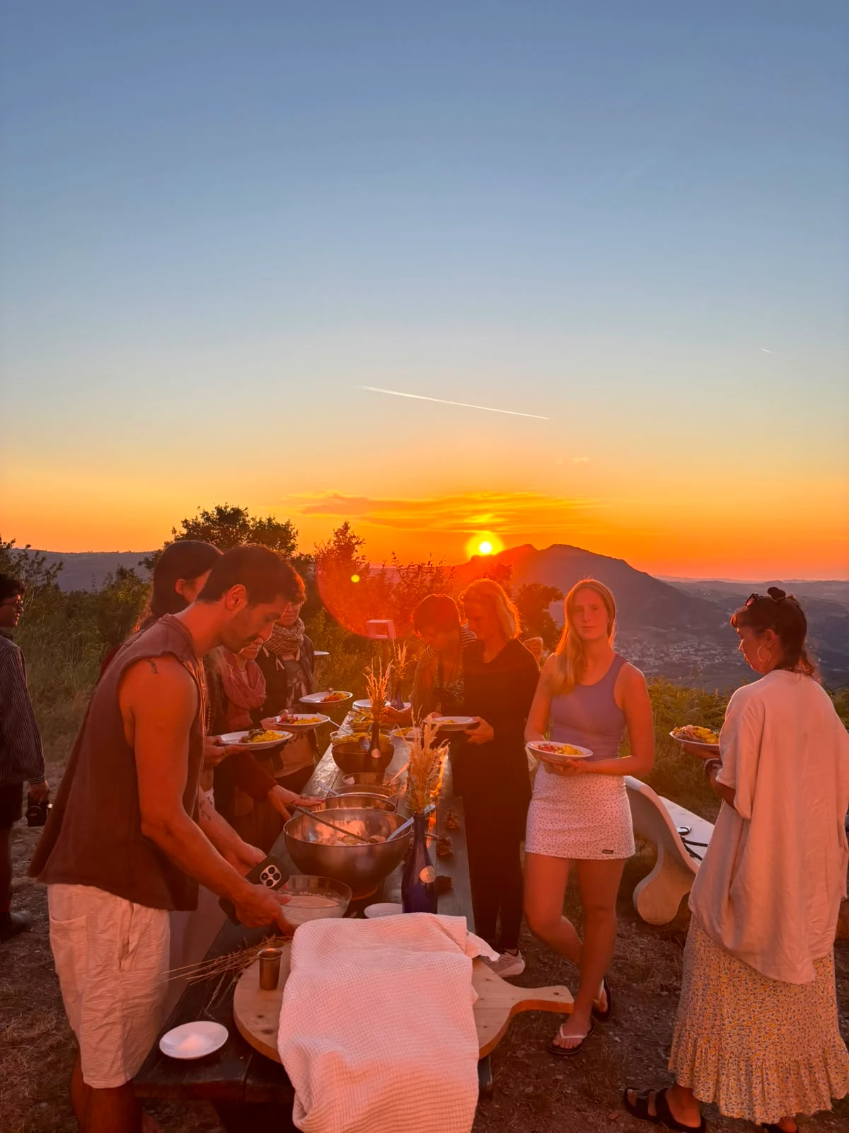 Community members sharing a long-table meal on a hilltop at Maiolo, sweeping valley and mountains glowing orange at dusk
