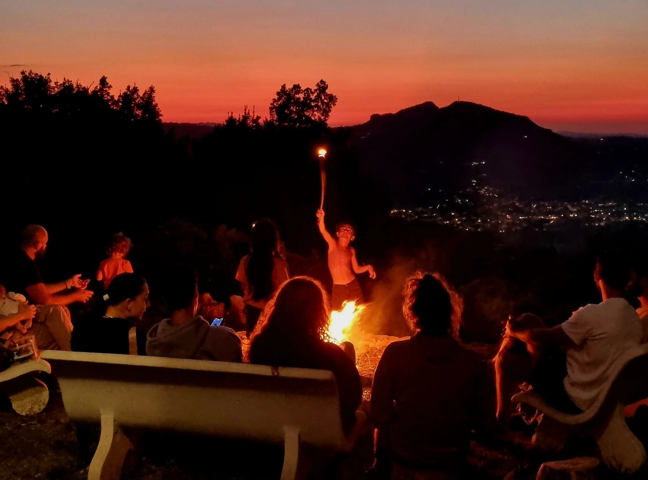 A group of people, including a child and a baby, gathered around a bonfire on a hilltop at dusk