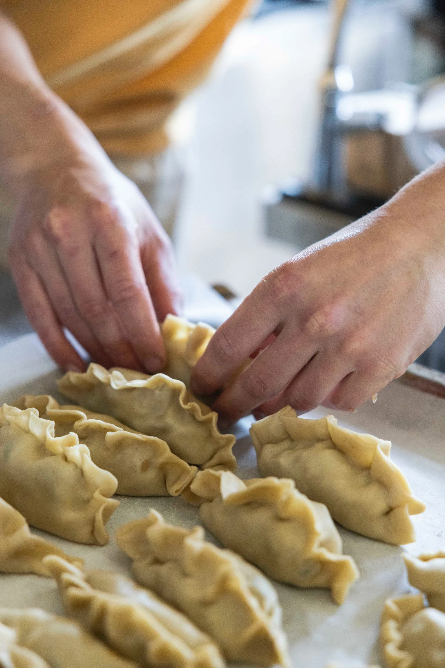Close-up of two pairs of hands shaping and crimping rows of dumplings on a floured countertop, a craft shared in the community kitchen