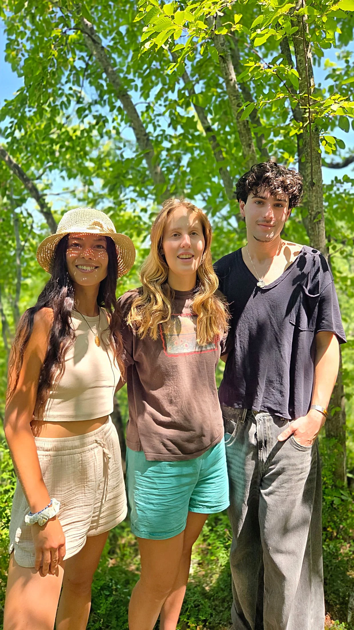 A young man and two young women smiling together under a leafy tree canopy